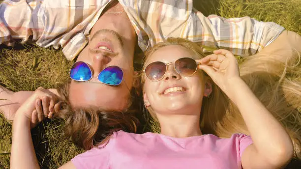 Happy young couple lying on lawn, holding hands, enjoying weekend in summer day