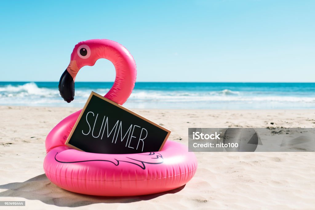 flamingo swim ring on the beach and text summer a signboard with the word summer written in it and a swim ring in the shape of a pink flamingo, on the sand of a beach, with the ocean in the background Summer Stock Photo