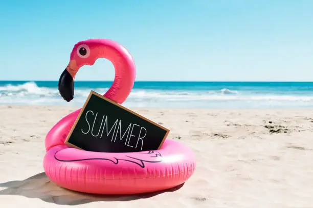 a signboard with the word summer written in it and a swim ring in the shape of a pink flamingo, on the sand of a beach, with the ocean in the background