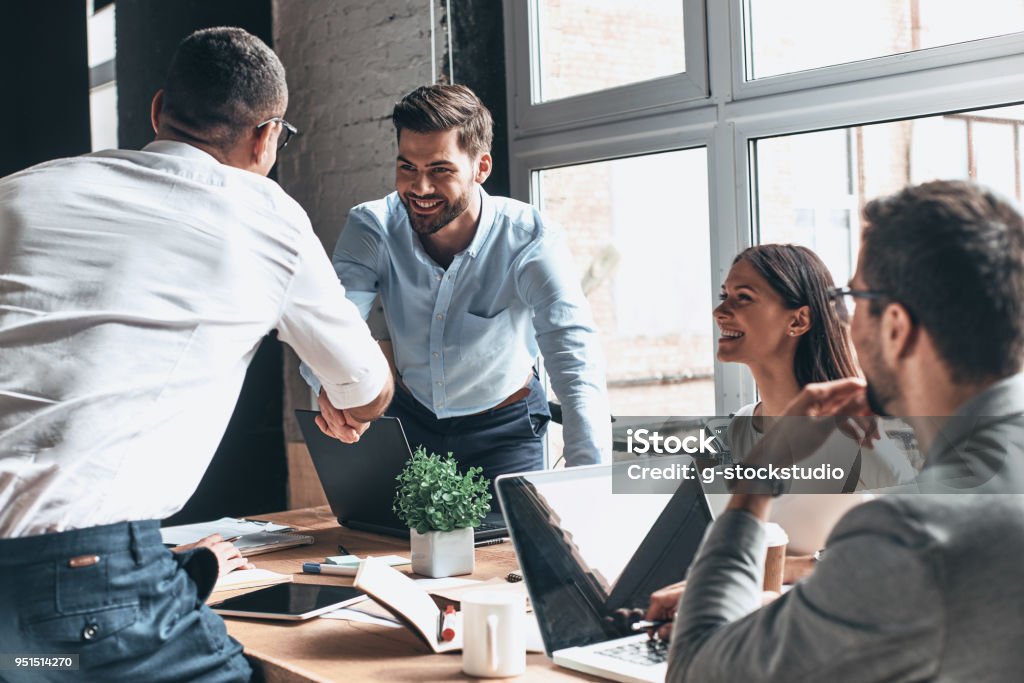 Glad to work with you! Young modern men in smart casual wear shaking hands and smiling while working in the creative office Business Stock Photo