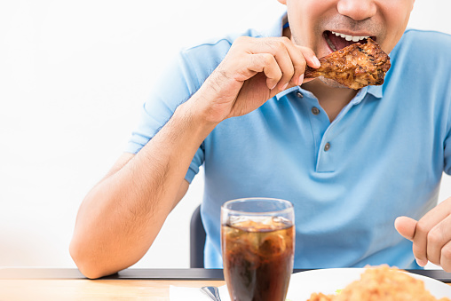 Young man happily eating grilled chicken drumstick with a soft drink at a restaurant.