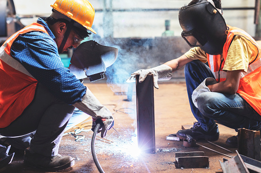Welder haphazardly holds his welding mask ahead of him while his juinor wears it properly. Two factory workers getting their tasks done.
