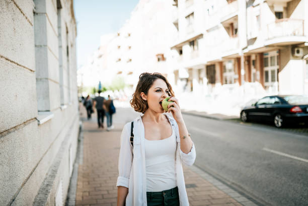 attraente giovane donna che cammina per strada e mangia la mela. - apple women green eating foto e immagini stock