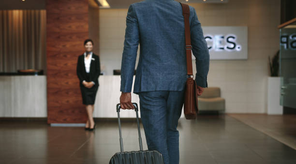 Businessman carrying suitcase while walking in hotel lobby. Business traveler arriving at hotel with female receptionist standing in background for welcoming.