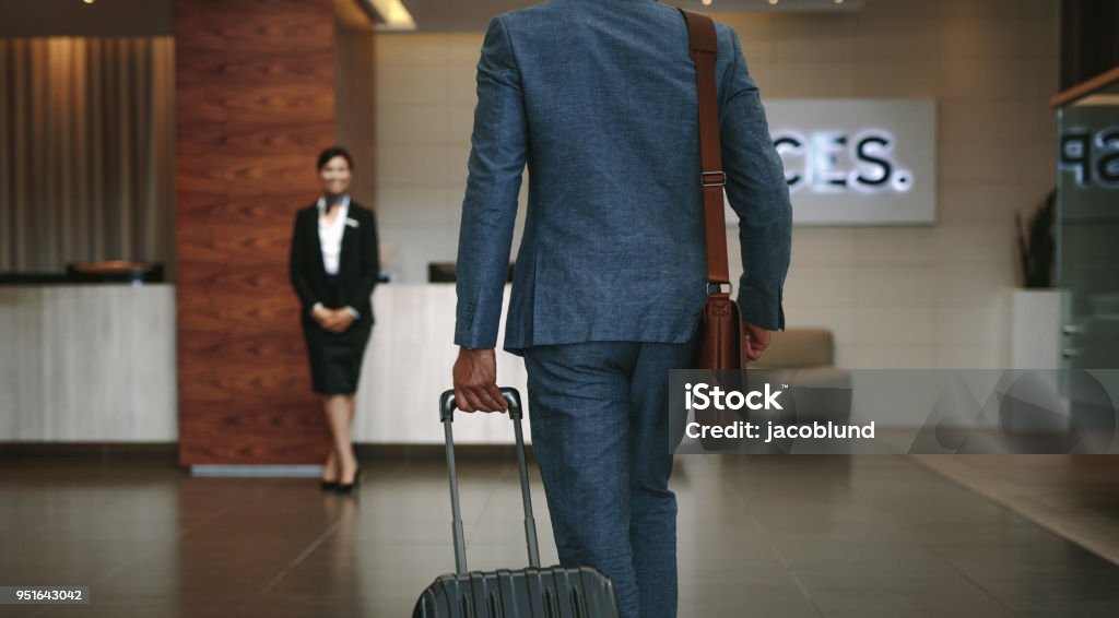 Business traveler arriving at hotel Businessman carrying suitcase while walking in hotel lobby. Business traveler arriving at hotel with female receptionist standing in background for welcoming. Hotel Stock Photo