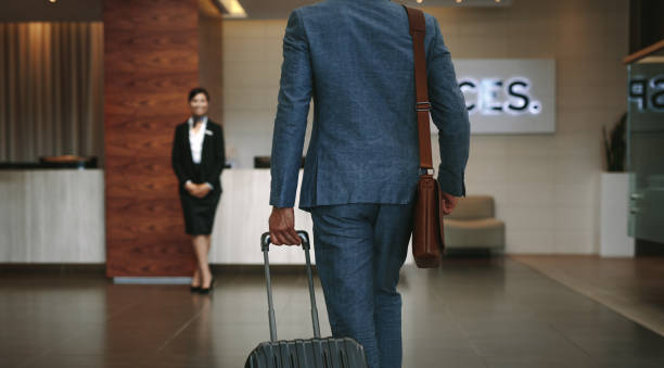 Business traveler arriving at hotel Businessman carrying suitcase while walking in hotel lobby. Business traveler arriving at hotel with female receptionist standing in background for welcoming. airport check in counter photos stock pictures, royalty-free photos & images