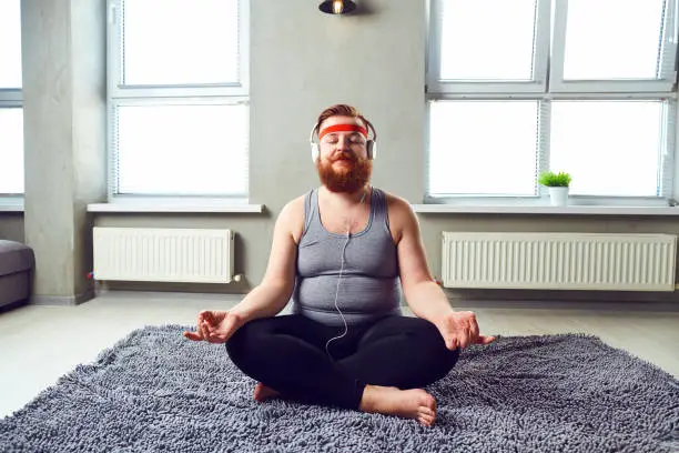 A funny fat bearded man in the headphones does yoga in the room.