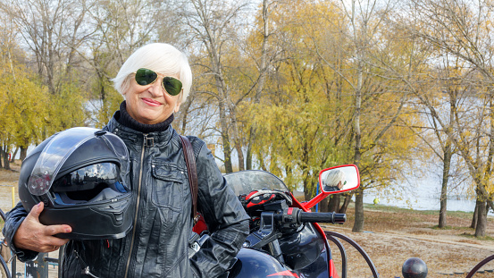 Portrait of a cheerful elderly woman with a motorcycle on the background of an autumn landscape