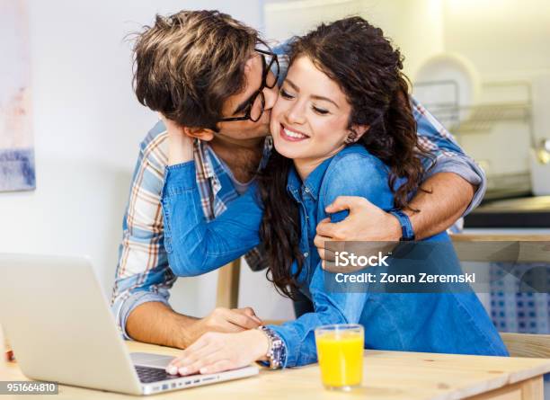 Young Couple Sitting By Table With Laptop And Eating Breakfast Early In The Morning Together Stock Photo - Download Image Now