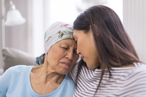 Asian elderly woman with cancer and wearing a headcovering is embracing her adult daughter. They are sitting on a couch and their foreheads are toughing.