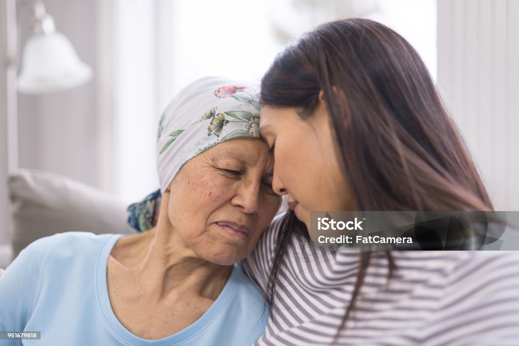 Ethnic elderly woman with cancer embracing her adult daughter Asian elderly woman with cancer and wearing a headcovering is embracing her adult daughter. They are sitting on a couch and their foreheads are toughing. Cancer - Illness Stock Photo