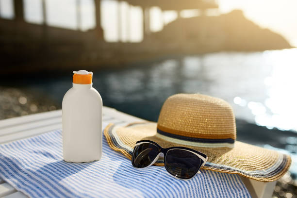 Suntan cream bottle and sunglasses on beach towel with sea shore on background. Sunscreen on deck chair outdoors on sunrise or sunset. Skin care and protection concept. Golden tan stock photo
