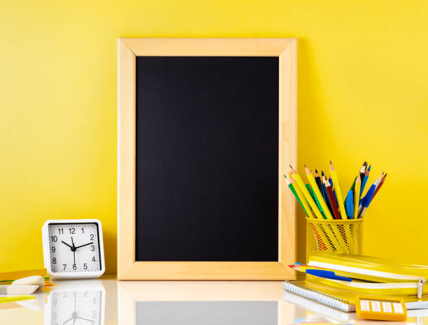 istock Chalkboard and school supplies on white table by the yellow wall. Side view, empty space for text. Back to school concept. 951703022