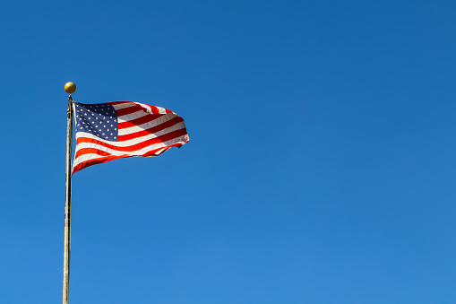 Horizontal full color view of the flags of the United States and the state if Hawaii rippling in the wind against a clear blue sky, with copy space. Both flags are red, white, and blue. While the U.S. flag has 50 stars in the top left corner, the Hawaiian flag has the British Union Jack.