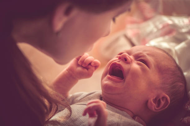 Mother holding crying baby girl wearing white dress lying on hands in room closeup