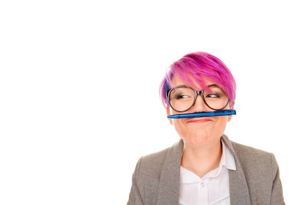 Adorable young millennial girl with pink hairstyle wearing glasses and holding pen between nose and lips having fun and looking away on white.