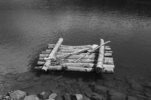 Black and white photo of a homemade wooden raft with a wooden ore tied  at the bank of a lake.