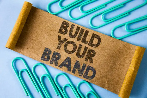 Photo of Text sign showing Build Your Brand. Conceptual photo create your own logo slogan Model Advertising E Marketing written on Folded Cardboard paper piece on plain blue background within Paper Pins.