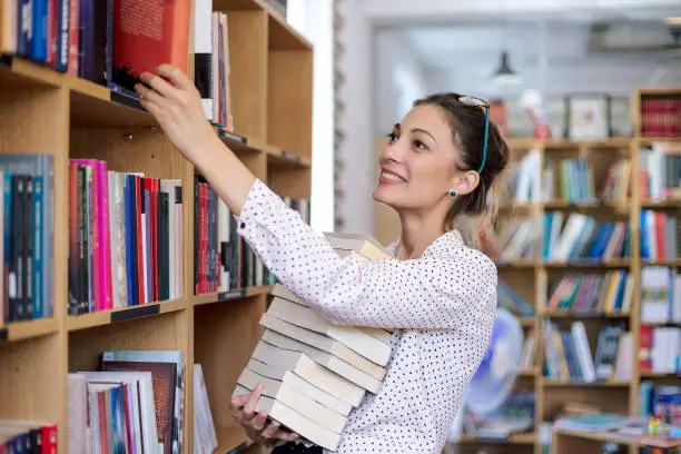 Attractive young woman wearing pink pale shirt with dots and fashionable glasses holding a stack of books in the library surrounded by bookshelves with colorful books.
