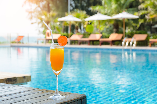 Orange juice with carrot slice in cocktail glass on wooden table at outdoor swimming pool, summer tropical holiday concept