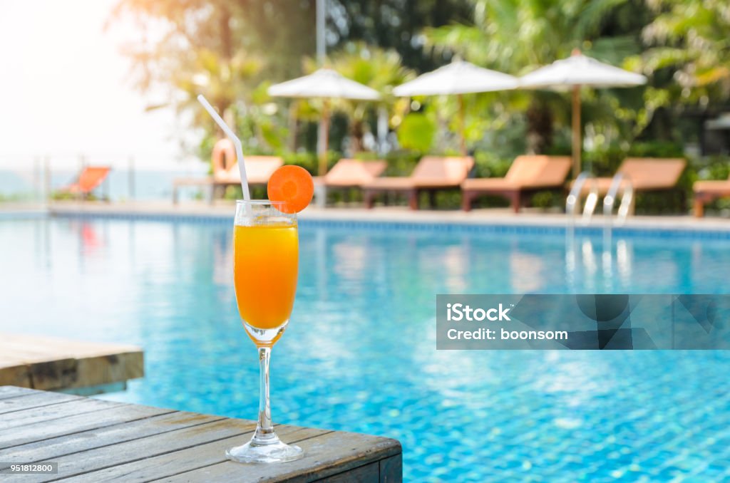 Orange juice glass on wooden table with swimming pool view background Orange juice with carrot slice in cocktail glass on wooden table at outdoor swimming pool, summer tropical holiday concept Swimming Pool Stock Photo