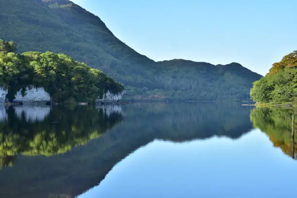 Reflections of hills forests and rock formations on calm surface of Muckross Lake in Ireland.