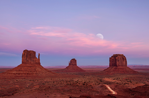 Long exposure on Valley Drive in Monument Valley showing the Mittens and cars travelling at the close of the park with the moon rising in the distance.