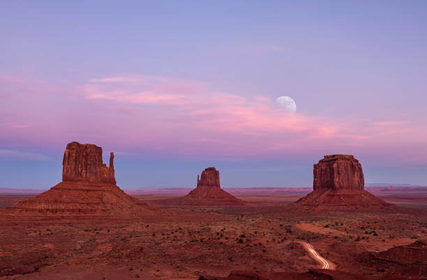 Sunset and Moon Rise at Monument Valley Long exposure on Valley Drive in Monument Valley showing the Mittens and cars travelling at the close of the park with the moon rising in the distance. monument valley photos stock pictures, royalty-free photos & images