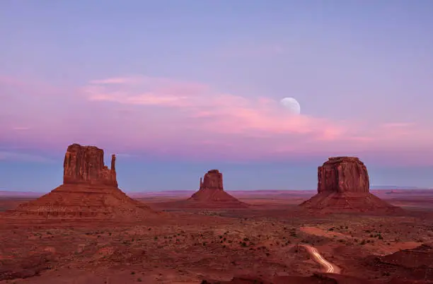 Long exposure on Valley Drive in Monument Valley showing the Mittens and cars travelling at the close of the park with the moon rising in the distance.