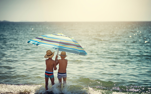 Funny kids are playing on a beach on a sunny summer day. Kids are standing in sea and holding a big beach umbrella.