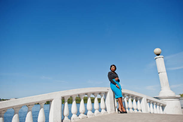 Stylish african american model girl in glasses, blue skirt and black blouse posed outdoor on bridge against lake.