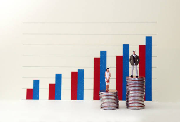A miniature man and woman standing on a pile of coins in front of a bar graph. The ongoing concept of the income gap between men and women. stock photo