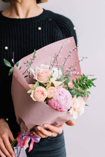 very nice young woman holding a colourful fresh blossoming flower bouquet of roses, carnations and peonies in pastel cream and pink colours on the grey wall background - flower head bouquet built structure carnation imagens e fotografias de stock