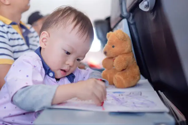 Little Asian 1 year old toddler boy coloring in coloring book with crayons during flight on airplane. Flying with children, Flight entertainment for kids, Happy air travel & little traveler concept