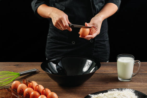 мужские руки шеф-повара разбивают яйцо на деревянном коричневом столе в черной миске. черный фон. концепция приготовления пищи. крупный пла - cake making mixing eggs стоковые фото и изображения