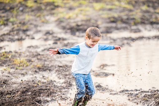 A young boy is outdoors on a summer day. He is having fun splashing in a mud puddle in a field.