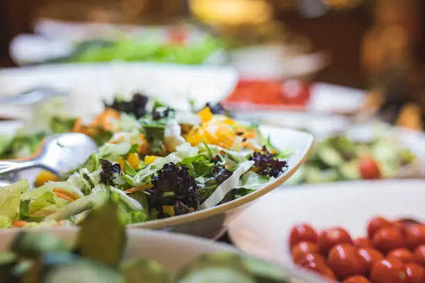 A fresh buffet of a colorful variety of healthy organic salads. Focus is on a salad bowl of mixed leafs and carrots, and there is a bowl of tomatoes on the right side.