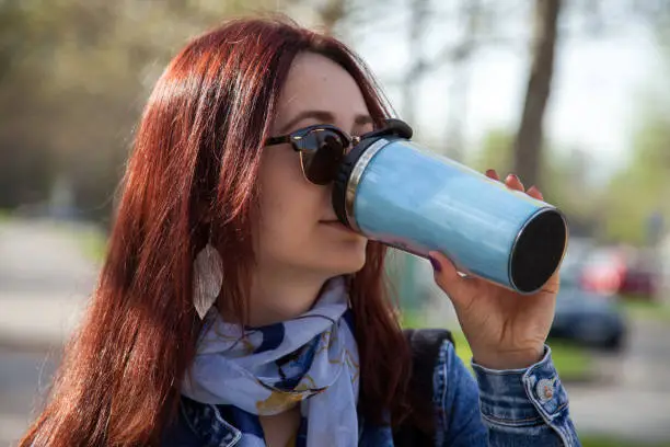 Close-up shot of a young woman drinking coffee from a travel mug in the city street.