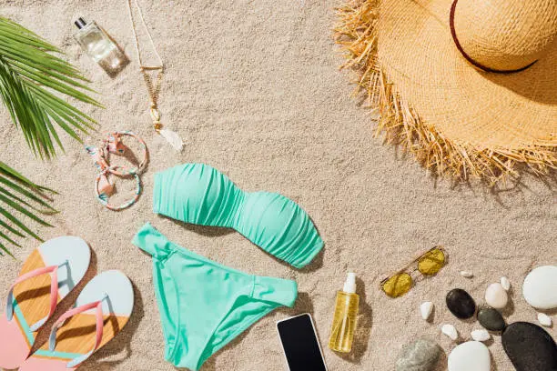 Photo of top view of blue bikini and flip flops with various accessories on sandy beach