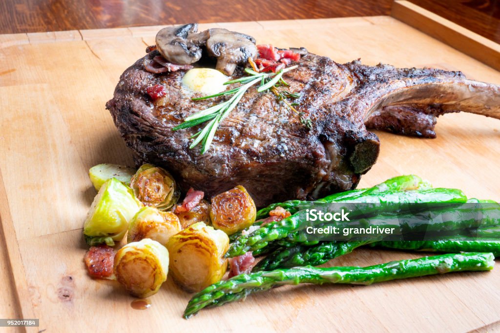 Huge Cowboy Cut Rib Eye Beef Steak with Ketogenic Vegetables on a Plate Large cowboy cut bone-in rib eye steak grilled up and on a plate with Brussels sprouts and Asparagus, a perfect ketogenic or low carb diet meal. Ketogenic Diet Stock Photo