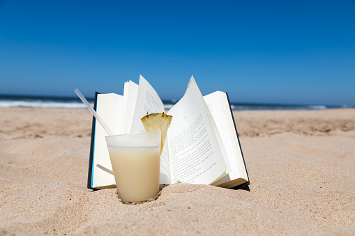 Pina Colada drink and an open book at the beach with a view of the ocean.