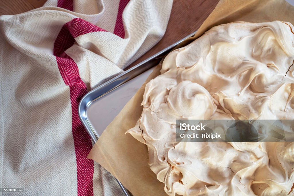 Making a Pavlova dessert Cooking the meringue base for a Pavlova dessert. Fresh meringue, with crisp golden top, on baking parchment and a stainless steel baking tray. Overhead view on wooden table with cloth. Meringue Stock Photo