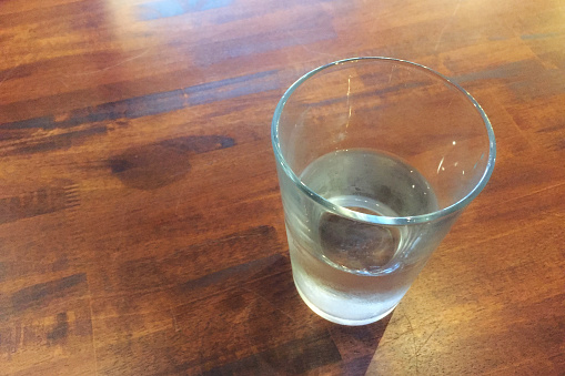 There is half water contain in a glass placed on the wood table at restaurant