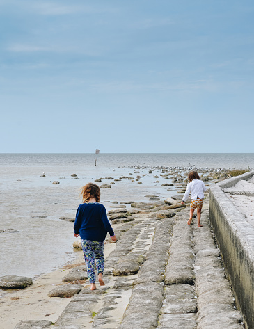 Two kids,  girls, walk along a seawall near the ocean, sandbags under their bare feet, beautiful body of water, coastline