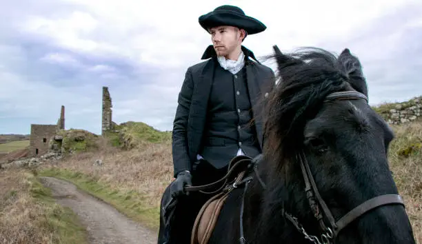 Good looking man, riding horse in Regency 18th Century Poldark Costume with stormy sky and tin mine ruins and Cornish countryside in background
