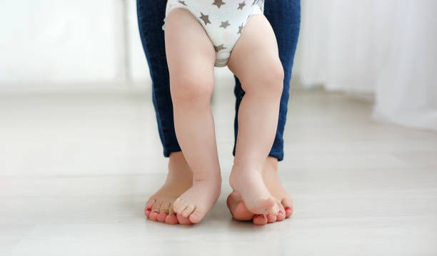 The bare feet of mother and baby on the floor in an empty room Bare legs of the child, dressed in a white diaper, standing on the bare soles of the feet of mother dressed in blue jeans, baby feet are between mothers legs, portrait on the background of the home environment. first steps stock pictures, royalty-free photos & images