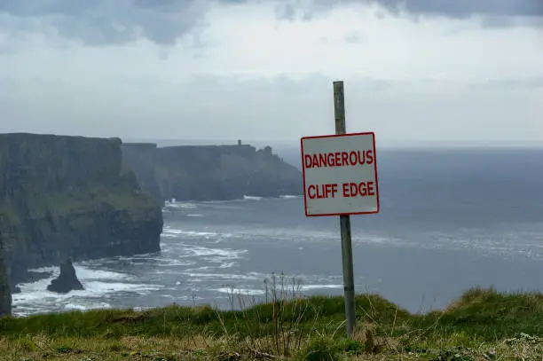 Photo of Dangerous cliff edge sign in overcast weather at Cliffs of Moher in Ireland. Danger sign warning of a vertical drop on coastal cliffs in typical irish weather.