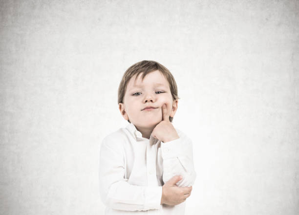 Cute little boy in a white shirt and dark blue jeans is thinking holding his finger near his cheek. Decision making concept. A concrete wall background.