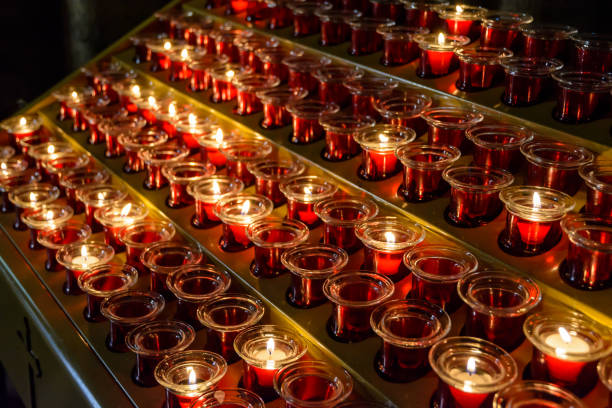 istock Small white votive candles are lighted and placed in red glass candlesticks among empty ones, lined up on a candle holder stand in a catholic church. 952111994