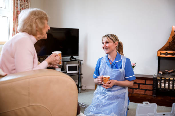 Home Visits For the Elderly A nurse sits on her knees after assisting an elderly lady. The senior woman is happy sitting in her armchair. They are both face to face chatting and enjoying a hot drink. english spoken stock pictures, royalty-free photos & images